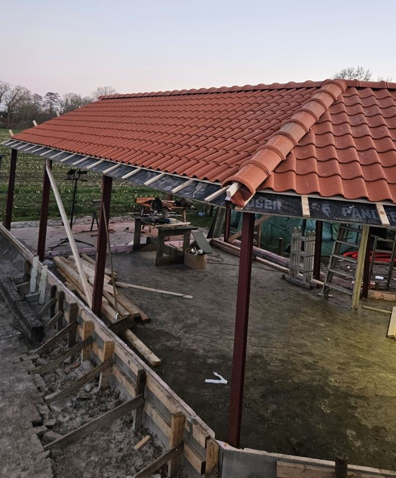 Bijgebouw met rode stalen kolommen en een oranje pannendak met hoekkeper, op een betonvloer